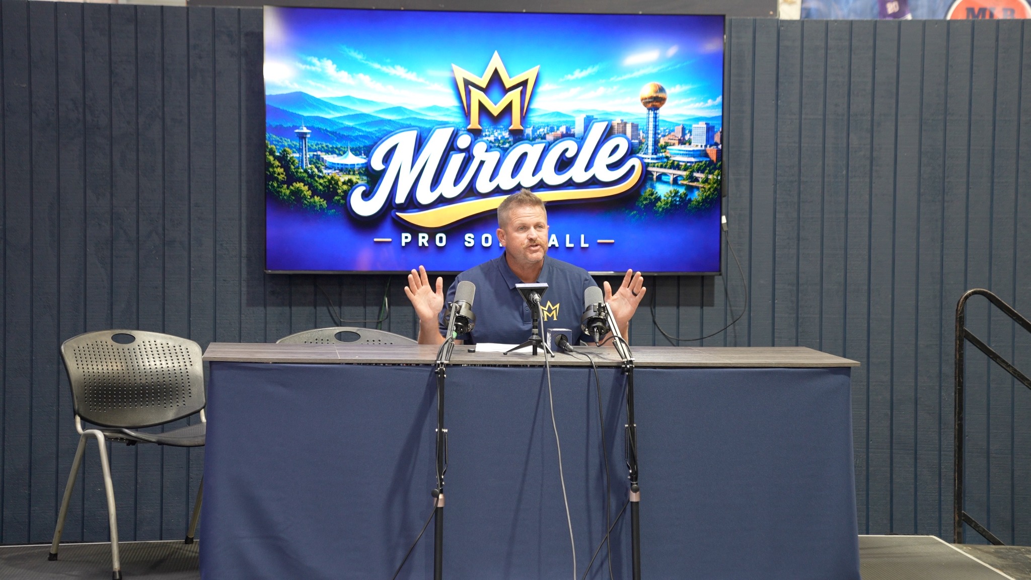 Eugene Bleecker speaking at a press table with the Knoxville Miracle screen behind him